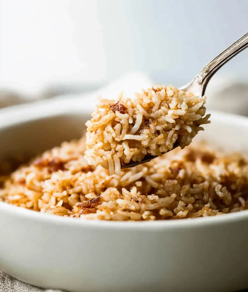 shows a bowl of "stick of butter rice," with a spoon lifting a portion of the moist, golden-brown rice. The dish has a rich, buttery sheen, highlighting its creamy texture, and is set against a softly lit background for a warm and inviting feel.