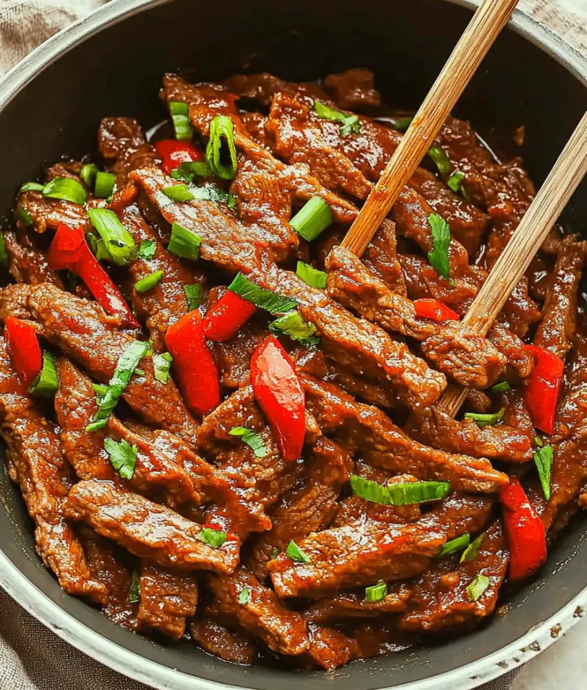 A pan of Beijing Beef featuring tender beef strips in a savory sauce, garnished with red bell peppers and green onions, with chopsticks resting on the side.