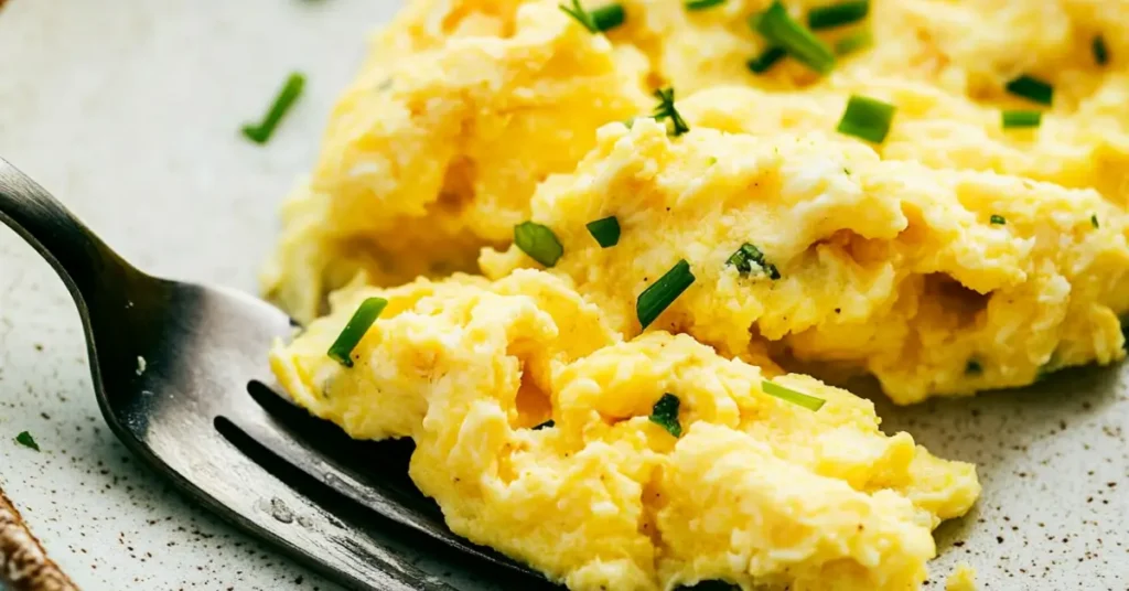 shows a close-up of fluffy scrambled eggs garnished with chopped chives, served on a plate with a fork. The eggs have a soft and creamy texture, perfectly baked for a golden finish.