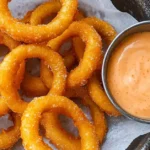 A close-up of golden crispy onion rings cooked in an air fryer, served with a bowl of creamy dipping sauce.