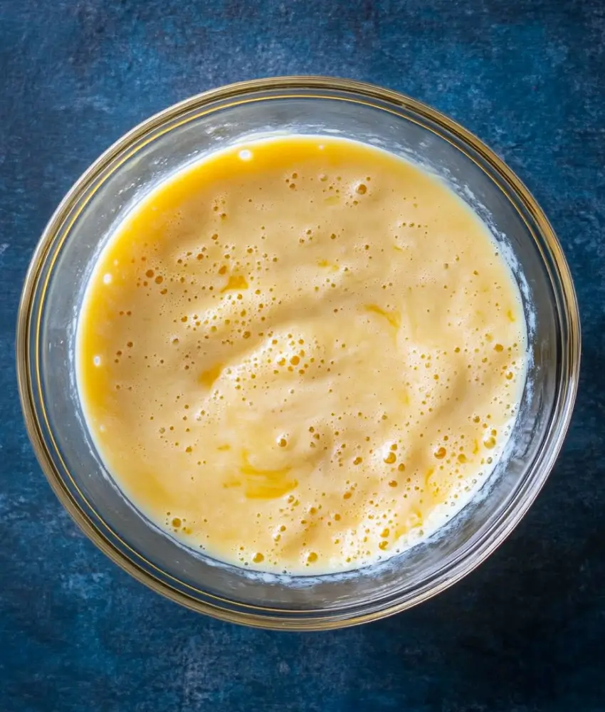 shows a glass bowl containing a smooth and frothy egg mixture, likely whisked and prepared for cooking or baking. The light yellow color and even texture indicate the eggs are well blended, ready for the next step in a recipe.