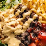 A close-up of a loaded burger bowl featuring ground beef, crispy French fries, fresh lettuce, diced tomatoes, and drizzled special sauce in a bowl.