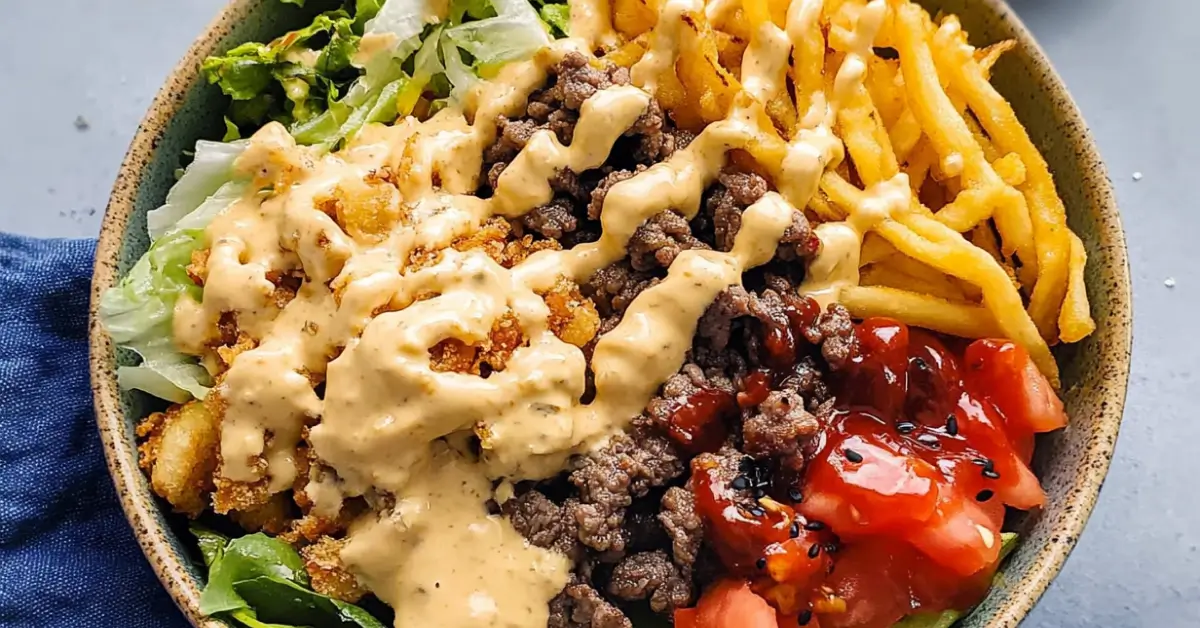 A close-up of a loaded burger bowl featuring ground beef, crispy French fries, fresh lettuce, diced tomatoes, and drizzled special sauce in a bowl.