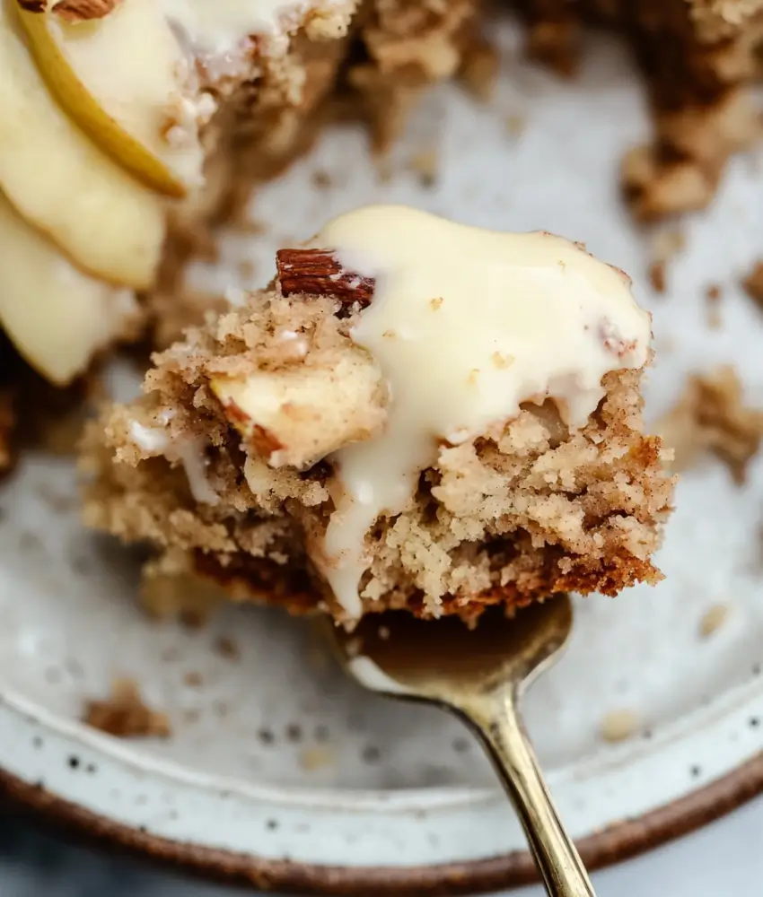 A close-up of a bite-sized piece of apple spice cake on a gold fork, featuring a moist, spiced crumb texture and creamy glaze. The cake piece is garnished with a chopped nut and appears soft and rich, with more cake and apple slices visible in the blurred background.