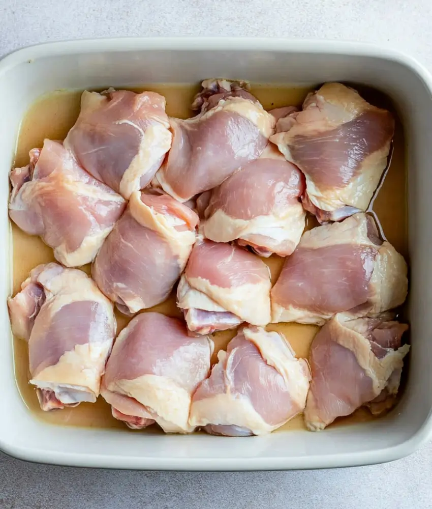 a baking dish filled with raw chicken thighs sitting in a light marinade or broth, preparing for a flavorful transformation into Huli Huli Chicken. The evenly arranged chicken pieces are ready to absorb the delicious marinade before cooking.