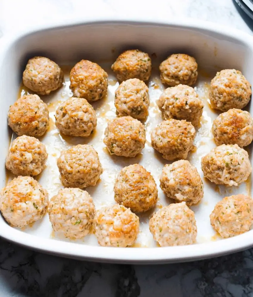 This image shows a baking dish filled with uncooked or partially cooked turkey meatballs, evenly spaced and lightly browned on some edges. The meatballs are sitting in a small amount of juices, indicating they are in the early stages of cooking or prepped for baking.