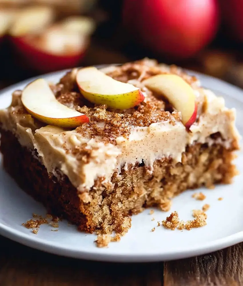 A slice of apple spice cake on a white plate, topped with creamy frosting, apple slices, and a sprinkle of cinnamon. The cake has a moist crumb and rich texture, with scattered crumbs on the plate and blurred red apples in the background.