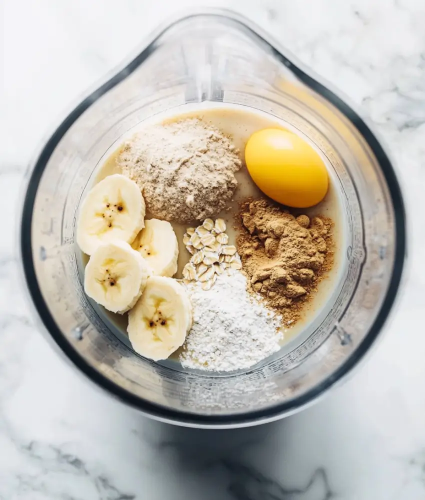 A close-up view of a blender filled with sliced bananas, oats, flour, an egg, cinnamon, and other pancake ingredients on a marble countertop.