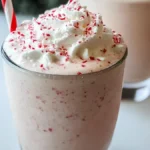 Close-up of a peppermint milkshake topped with whipped cream and crushed peppermint candies in a clear glass with a festive red and white straw.