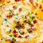 Close-up of crack chicken dip topped with melted cheese, crispy bacon bits, and green onions in a white baking dish, surrounded by tortilla chips.