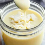 A glass jar filled with creamy white chocolate syrup, topped with white chocolate shavings, with a spoon drizzling syrup into the jar.