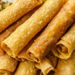 A plate of golden, crispy chicken taquitos served with diced tomatoes and jalapeños in the background.