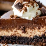 A slice of brownie cheesecake with a chocolate crust, creamy cheesecake center, and rich chocolate ganache topping, garnished with whipped cream, chocolate shavings, and cocoa powder.