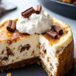 A slice of cinnamon roll cheesecake topped with whipped cream and cinnamon sticks, served on a dark plate with a creamy filling and a golden-brown crust.