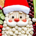 A colorful Santa-themed veggie tray featuring cauliflower, cherry tomatoes, cucumbers, celery, olives, and crackers arranged to resemble Santa Claus wearing a red hat.