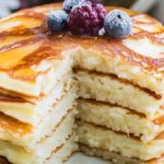 A stack of fluffy buttermilk pancakes topped with blueberries, blackberries, and a drizzle of golden maple syrup, served on a rustic plate.