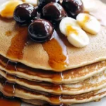 A stack of golden brown pancakes topped with sliced bananas, dark cherries, and drizzled with maple syrup on a white plate.