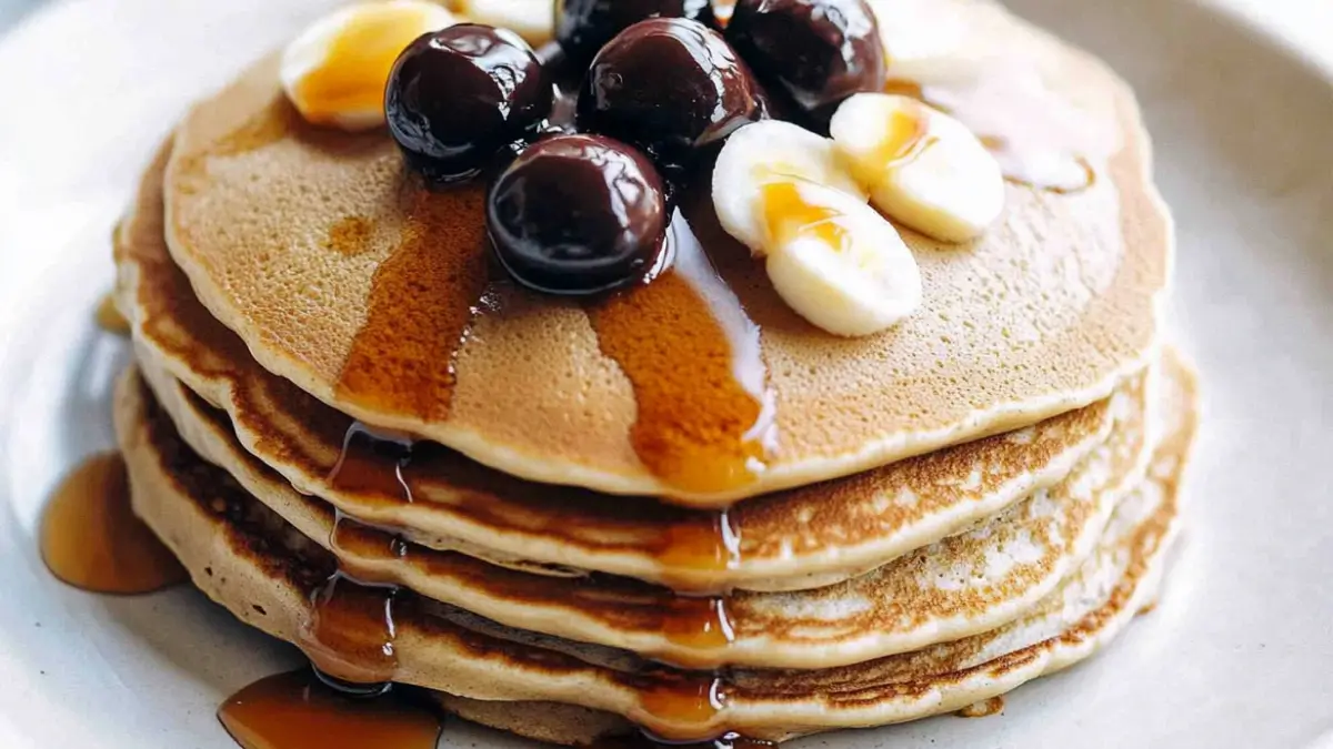 A stack of golden brown pancakes topped with sliced bananas, dark cherries, and drizzled with maple syrup on a white plate.