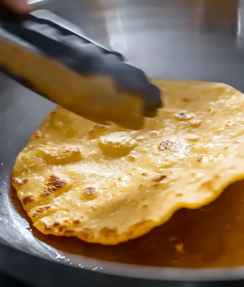 A close-up of a golden tortilla being cooked in a pan, lifted by tongs, with visible bubbles and charred spots.