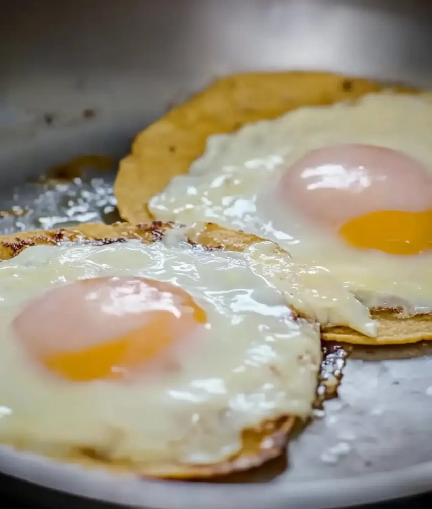 Close-up of two sunny-side-up eggs cooking on crispy tortillas in a pan, with golden edges and glossy whites.