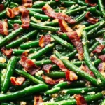 Sautéed green beans with crispy bacon pieces in a black skillet, topped with garlic and seasoning.