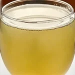 A glass mug filled with warm green tea, with a slice of lemon in the background