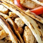 Three grilled chicken pitas filled with chicken, red onions, cucumbers, and tomatoes, served with a small bowl of tzatziki sauce on the side.