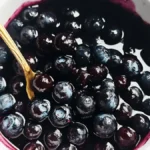 Close-up of a bowl of homemade blueberry sauce with a golden spoon, showing glistening blueberries in a rich, dark syrup.