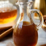 A glass pitcher filled with rich brown homemade cinnamon syrup surrounded by cinnamon sticks and a blurred background featuring jars and warm lighting.