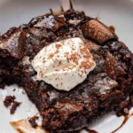 A close-up of a gooey chocolate cobbler topped with whipped cream and dusted with cocoa powder, served in a white bowl.