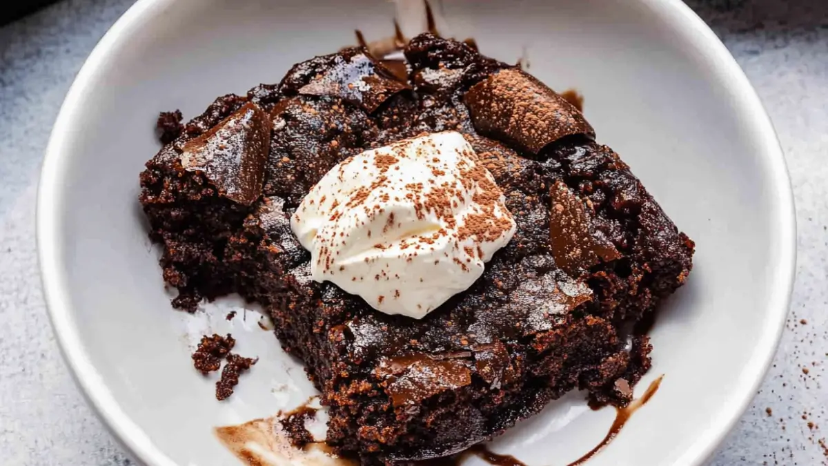 A close-up of a gooey chocolate cobbler topped with whipped cream and dusted with cocoa powder, served in a white bowl.