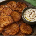 Crispy spiral onion ring chips served on a dark plate with a bowl of creamy white dip topped with red pepper flakes