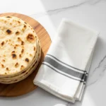 A stack of warm, golden-brown pita bread with subtle char marks, piled on a round wooden cutting board. A folded white kitchen towel with gray stripes rests beside it on a light marble counter.